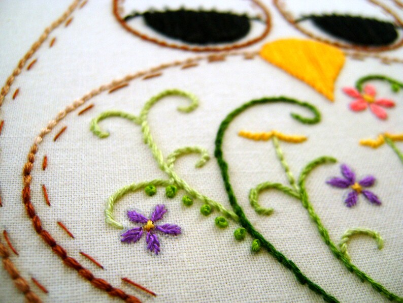May include: Close-up of a white fabric with embroidered details. The design features an owl with black eyes, a yellow beak, and floral accents in green, purple, and pink. The stitching is in various colors, including brown and orange.