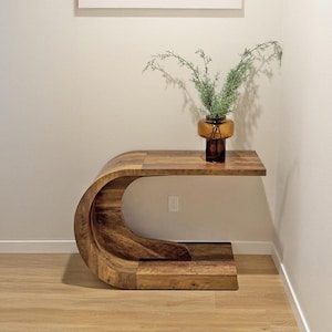 May include: A wooden console table with a unique curved design. The table has a natural wood finish and a shelf. A brown glass vase with greenery sits on the table. A framed abstract art piece hangs above the table.