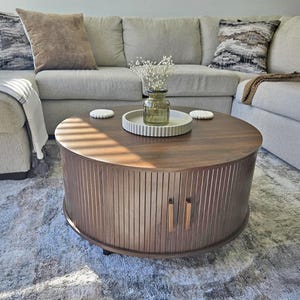 May include: A round, dark brown coffee table with vertical wood paneling and two small, vertical handles. The table sits on a light gray rug in front of a light gray sectional sofa with decorative pillows and a vase of flowers.