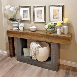 Op de afbeelding: Een houten console met een grijze basis en een bruin blad. De tafel is versierd met ingelijste kunstwerken, kaarsen, een decoratieve kom en een grote witte vaas. Een witte geweven mand en een crèmekleurige deken liggen op de onderste plank.