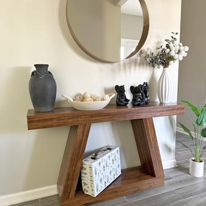 Pode incluir: Uma mesa de console de madeira escura com uma prateleira inferior e um tampo decorativo. O tampo exibe um vaso preto, uma tigela branca com esferas decorativas e três pequenas figuras de elefantes pretos. Um espelho redondo está pendurado acima.