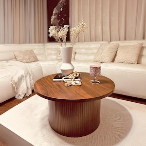 May include: A round, dark wood coffee table with a cylindrical base sits on a patterned rug. A white vase with dried flowers, a decorative candle, and a stack of books are on the table. A white sectional sofa and artwork are in the background.