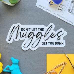Può includere: Un adesivo con la scritta "DON'T LET THE MUGGLES GET YOU DOWN" in corsivo nero. L'immagine include anche una statuina di mucca blu, una spilla blu e gialla, un biglietto e altri elementi decorativi su una superficie grigia.