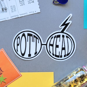 Pode incluir: Um adesivo branco com as palavras "POTT HEAD" em um design circular que se assemelha a óculos. Um raio está acima da palavra "HEAD". Outros itens incluem um bilhete, um alfinete de uma xícara azul e um frasco cheio de glitter dourado.