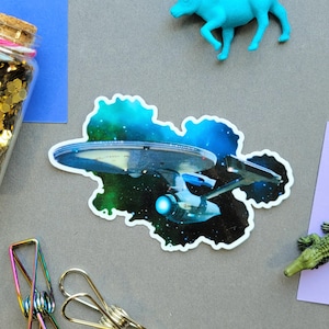 May include: A sticker featuring the Starship Enterprise from Star Trek, set against a nebula background. The image is surrounded by a variety of objects, including a bottle filled with gold glitter, paper clips, and toy animals.