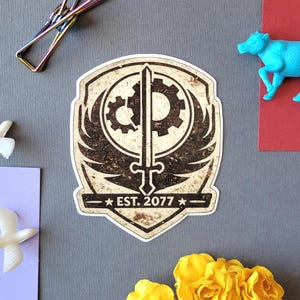 May include: A distressed sticker with a shield design, sword, gears, and wings. The text "EST. 2077" is on a banner. Decorative elements include a blue cow figurine and white dove figurines. The sticker has a vintage aesthetic.