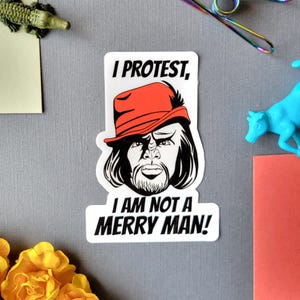 Puede incluir: Una pegatina blanca con el texto "I PROTEST, I AM NOT A MERRY MAN!" presenta una ilustración en blanco y negro de una persona con un sombrero rojo. La pegatina está sobre una superficie gris con otros objetos pequeños.