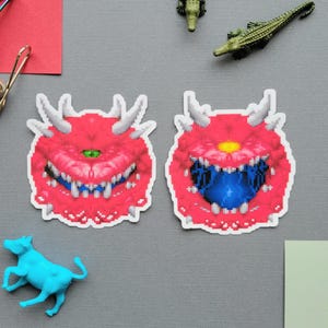 Könnte beinhalten: Zwei rote Pixel-Art-Aufkleber eines Monsters mit Hörnern, Zähnen und einem einzelnen Auge. Ein blaues Spielzeugrind, ein grünes Spielzeugkrokodil und Büroklammern sind ebenfalls auf einer grauen Oberfläche zu sehen.