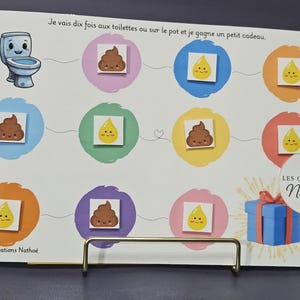 May include: A white board with a cartoon toilet, poop, and pee drop graphics. The board has the text "Je vais dix fois aux toilettes ou sur le pot et je gagne un petit cadeau." A blue gift box with a red ribbon is also shown.