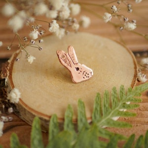 Puede incluir: Un pin de conejo de color cobre con una superficie texturizada. El pin está sobre una superficie de madera con una planta verde en primer plano.