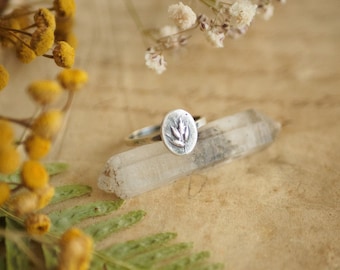 May include: A silver ring with a floral design on a white crystal. The ring is on a piece of parchment paper with dried flowers.