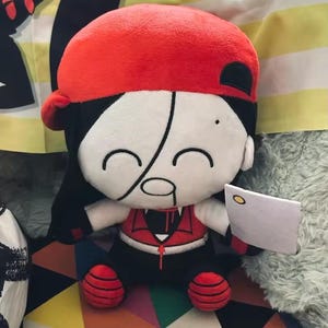 Puede incluir: Muñeco de peluche con gorra roja, pelo negro y cara blanca con una línea negra en el medio. El muñeco lleva un chaleco rojo y pantalones negros, y sostiene un pequeño rectángulo blanco. El muñeco está sobre una superficie estampada colorida.