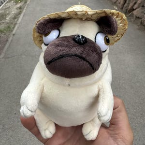 May include: A cream-coloured pug soft toy wearing a straw hat. The pug has a brown snout and large eyes. The hat is made of woven straw. The toy is held in a person's hand.