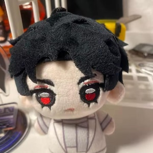 Puede incluir: Un muñeco de peluche con cabello negro y grandes ojos con borde rojo. El muñeco tiene un atuendo blanco con detalles grises. La cara del muñeco tiene maquillaje de ojos rojo y una boca pequeña. Es un artículo de colección.