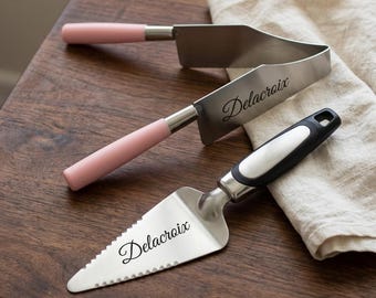 Könnte beinhalten: Ein Tortenheber aus Edelstahl mit schwarzem Griff und dreieckiger gezahnter Klinge, kombiniert mit einem Tortenmesser aus Edelstahl mit zwei hellrosa Griffen und dem eingravierten Text Delacroix auf beiden Metallflächen.