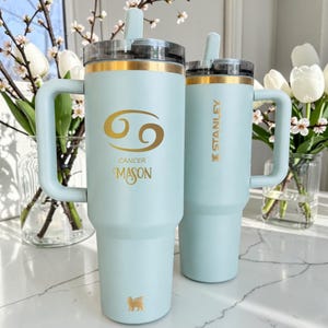 Könnte beinhalten: Zwei hellblaue Isolierbecher mit Griffen und durchsichtigen Deckeln. Der größere Becher zeigt ein goldenes Krebs-Sternzeichen, den goldenen Schriftzug "CANCER" und "MASON" sowie ein kleines goldenes Logo. Der kleinere Becher trägt das goldene "STANLEY"-Logo.