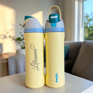 May include: Two pale yellow water bottles with teal and light blue accents. The left bottle features the name Lilianna in cursive script with a heart icon. Both bottles have a push-button lid mechanism with a carry loop and the Owala brand logo.
