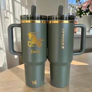 Könnte beinhalten: Zwei olivgrüne Edelstahlbecher mit Griffen und Trinkhalmen. Der linke Becher zeigt ein goldenes Steinbock-Sternzeichen und den Namen Emily. Der rechte Becher zeigt das goldene Stanley-Markenlogo vertikal an der Seite angebracht.