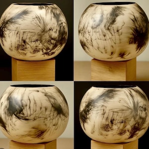 May include: Four images of a round, cream-colored wooden bowl with a dark, abstract design. The bowls have a black interior and sit on a small, square wooden base. The design resembles a mix of brushstrokes and natural patterns.