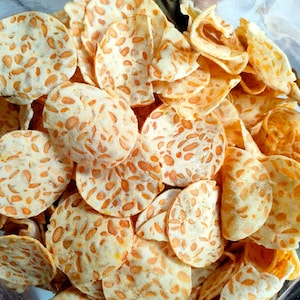 Könnte beinhalten: Eine Ansammlung dünner, runder und knuspriger Tempeh-Chips. Jeder Chip ist hellbeige und weist auf der gesamten Oberfläche sichtbare, eingebettete orange-braune Sojabohnenstücke auf, was dem Snackhaufen ein gesprenkeltes Aussehen verleiht.