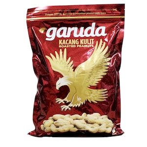 Op de afbeelding: Een glanzende rode snackzak met een grote gouden adelaarsafbeelding, de tekst garuda KACANG KULIT ROASTED PEANUTS en een hoopje lichtbruine pinda's in de dop onderaan. De bovenkant van de zak bevat een ritssluiting en extra tekst.