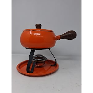 May include: A bright orange fondue pot with a matching lid and a dark brown wooden handle. The pot sits on a black metal stand with a circular orange base and includes a small metal burner component positioned centrally beneath the pot bowl.