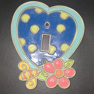 May include: Decorative light switch cover in the shape of a heart. The blue heart has yellow polka dots and a light blue border. A pink flower and a yellow and orange bee are on the bottom.