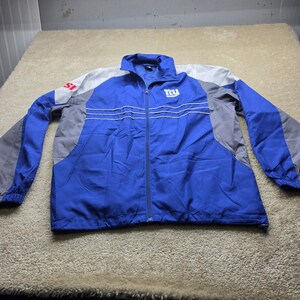 May include: A blue, gray, and white zip-up track jacket featuring the letters NY in white on the upper left chest and the letters SI in red on the right shoulder. The jacket has a high collar, long sleeves, and thin white horizontal stripes.