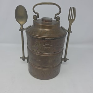 May include: A vintage brass tiffin carrier with a handle, spoon, and fork. The three-tiered container features etched floral designs. The spoon and fork are attached to the sides of the carrier. The handle is arched.