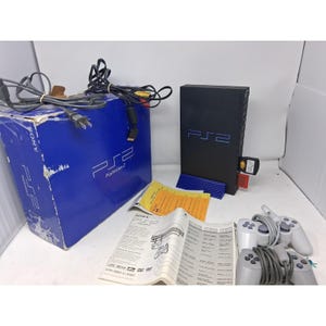 May include: A black Sony PlayStation 2 console standing vertically on a blue base, accompanied by a blue retail box, two grey wired controllers, a printed instruction manual, and yellow informational inserts, all featuring the PS2 logo.