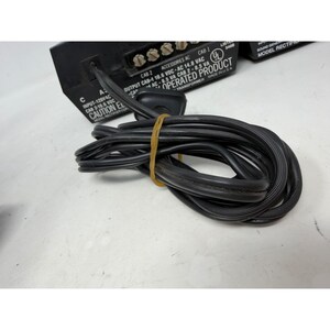 May include: A black electrical transformer with a coiled black power cord secured by a yellow rubber band. The device features white printed technical specifications including input and output voltage ratings, terminal screws, and a UL certification mark.