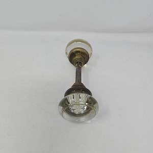 May include: Antique glass door knob with a clear, faceted design. The knob has a brass-colored base and a metal rod connecting to a smaller, rounded glass knob. A vintage hardware piece.