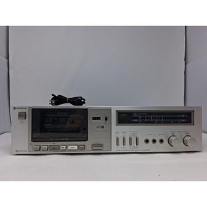 May include: A silver Hitachi D-E20 stereo cassette tape deck featuring a front-loading cassette compartment, soft-touch control buttons, a digital peak meter display, multiple toggle switches, two circular knobs, and a black power cord.