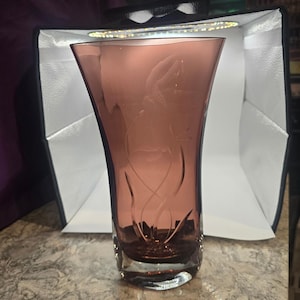 May include: A tall, mauve-colored glass vase with a flared top and a clear, etched floral design. The vase has a thick base and a smooth, polished finish, suitable for displaying flowers or as a decorative piece.