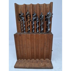 May include: A wooden storage case containing a set of metal auger drill bits. The case features vertical slots for each bit, with handwritten labels such as No. 16, 15, 14, 13, and 12 visible at the top. The metal bits have spiral flutes and pointed tips.