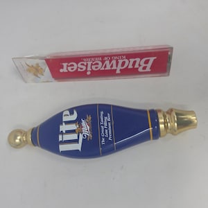 May include: Two beer tap handles. One is a blue Miller Lite tap handle with gold accents and text. The other is a Budweiser tap handle with red and white text and a clear plastic casing.