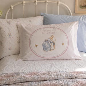 Puede incluir: Una cama con ropa de cama y almohadas blancas. La almohada central presenta una ilustración de Peter Rabbit con el texto "THE TALE OF PETER RABBIT". Otras almohadas tienen estampados florales y azul claro liso. Una imagen enmarcada cuelga en la pared.