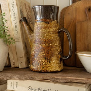 May include: A ceramic pitcher with a dark brown glazed rim and handle, featuring a textured, speckled mustard yellow and brown body. The pitcher has a tapered spout and a sturdy handle, standing upright with a glossy finish on the upper section.