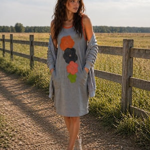 Op de afbeelding: Een grijze mouwloze jurk met een bloemenontwerp, met oranje, zwarte, rode en groene bloemen. De jurk heeft een rode bies en zakken. Een grijze cardigan is over de schouders gedrapeerd. De persoon draagt grijze sneakers en loopt op een zandpad.