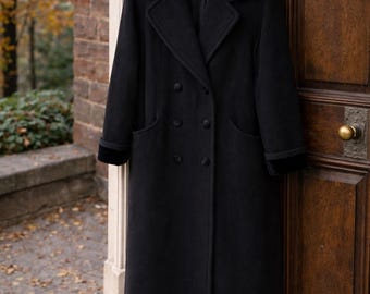 May include: A long, black wool coat with a velvet collar and cuffs hangs on a wooden hanger. The double-breasted coat has two rows of buttons and two front pockets. The coat is hanging on a wooden door.
