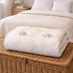 May include: A folded white quilt featuring a stitched circular pattern across the surface. The quilt is decorated with small, pastel-colored floral appliques in pink and blue, along with square, cream-colored crocheted accents placed throughout.
