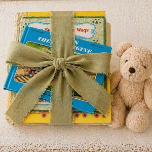 Könnte beinhalten: Ein Stapel Kinderbücher, gebunden mit einem salbeigrünen Band und einer Schleife, neben einem Teddybär. Die Bücher haben blaue und gelbe Einbände mit Illustrationen und Text. Der Teddybär ist hellbraun.