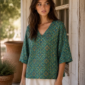 May include: A teal V-neck blouse featuring a repeating intricate geometric pattern in shades of gold, orange, and dark blue. The top has loose, three-quarter length sleeves and a relaxed fit, paired with plain off-white trousers.