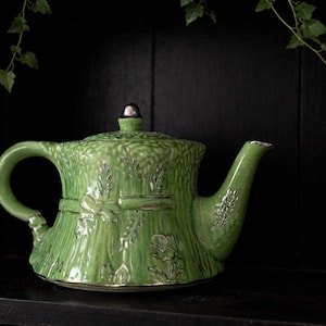 May include: A green ceramic teapot with a textured wheat stalk design. The teapot has a silver-colored lid and spout trim. The handle and body are adorned with embossed floral and wheat motifs. The teapot is displayed on a black shelf with a dark background.