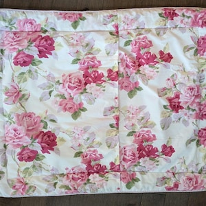 May include: A rectangular, floral-patterned quilt featuring a cream background with a repeating pattern of pink and red roses and green leaves. The quilt has a ruffled edge and is likely made of cotton or a similar fabric. The overall aesthetic is vintage and romantic.