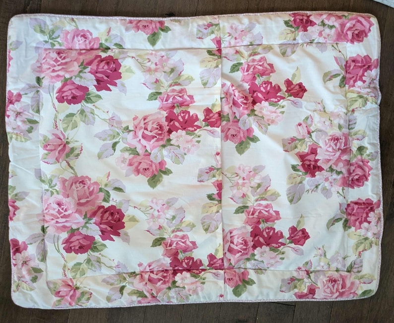 May include: A rectangular, floral-patterned quilt featuring a cream background with a repeating pattern of pink and red roses and green leaves. The quilt has a ruffled edge and is likely made of cotton or a similar fabric. The overall aesthetic is vintage and romantic.