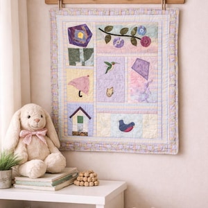 May include: A pastel-colored patchwork quilt with various appliqued shapes, including a house, umbrella, and bird, hangs on a wall. A plush bunny with a pink bow sits on a white table next to books and a woven basket.