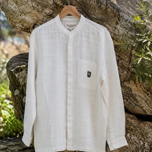 May include: A long-sleeved, off-white shirt with a mandarin collar and a single chest pocket. The shirt features a small, dark rectangular patch on the pocket with a graphic design and the text FIVE COLOURS EARTH on the inner neck label.