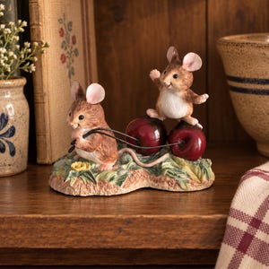 May include: A decorative figurine featuring two brown and white mice interacting with two large, glossy red cherries. One mouse stands on the cherries while the other pulls them by their stems. The base is textured to resemble green foliage.