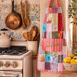 May include: A colourful patchwork apron with a pink pocket embroidered with the word "Pastiamo" hangs on a wall. The apron features a variety of fabric patterns and colours, including floral, polka dots, and plaid. A vintage kitchen scene with copper pots and a white stove is in the background.